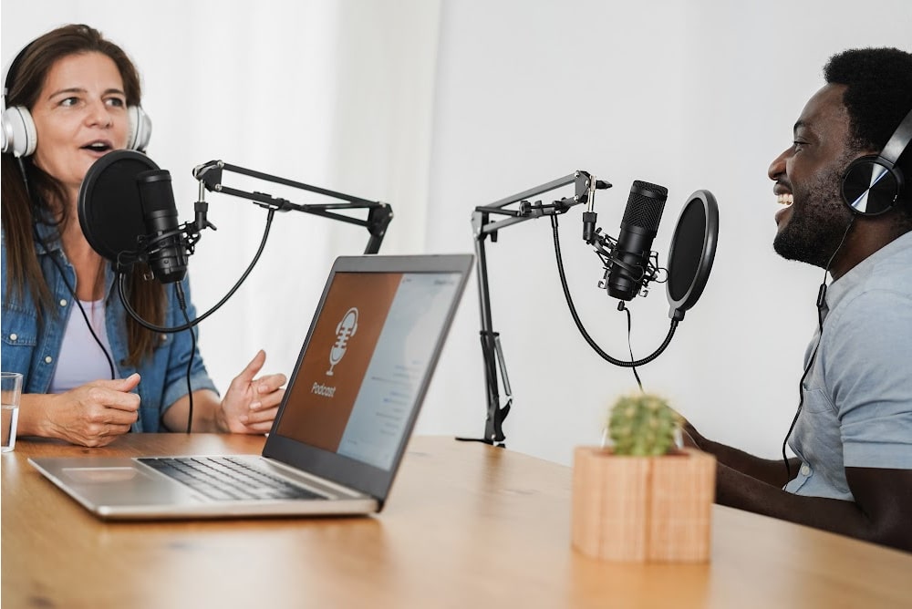 woman over 40 and a man with microphones and a laptop recording a podcast