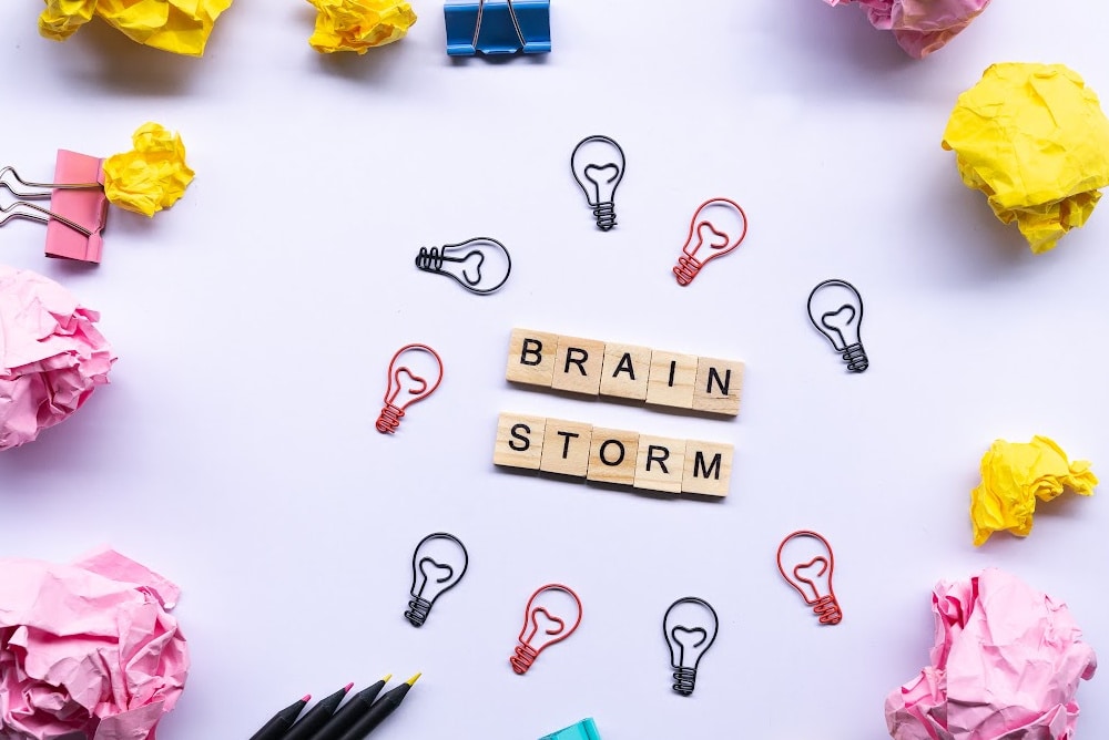 the word brainstorm in scrabble tiles lightbulbs paper clips and balled up paper