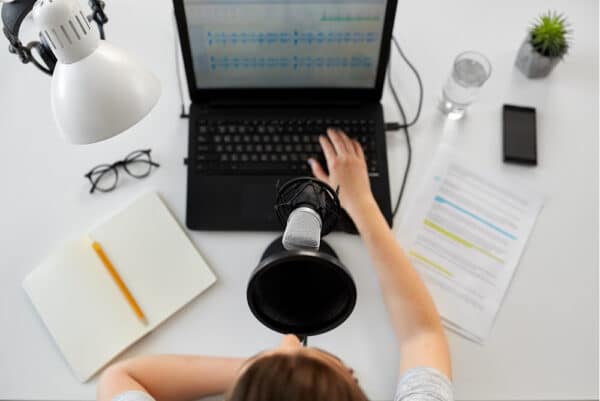 overhead view of a desk with a laptop mic notebook script light glasses and a person with there hand on the laptop to post your podcast